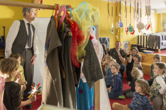 Mann steht mit zwei Kleinkindern hinter Bettlaken, vor dem Bettlaken sitzt eine Gruppe von Kleinkindern