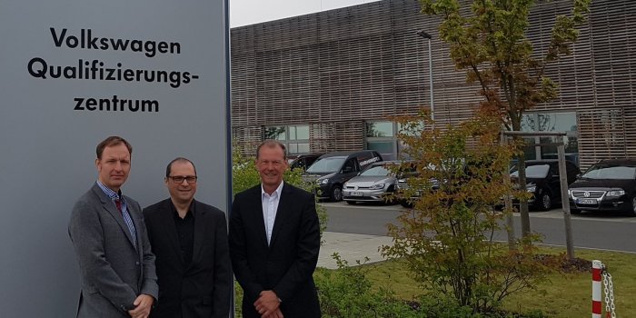 3 Männer stehen vor einem Pylon mit der Aufschrift "Volkswagen Qualifizierungszentrum". 