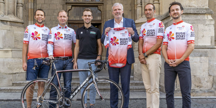 Sechs Männer stehen mit einem Rad und in Sportkleidung vor einem alten Gebäude.