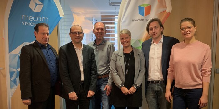 6 Personen stehen vor Werbeaufstellern von mecom. 