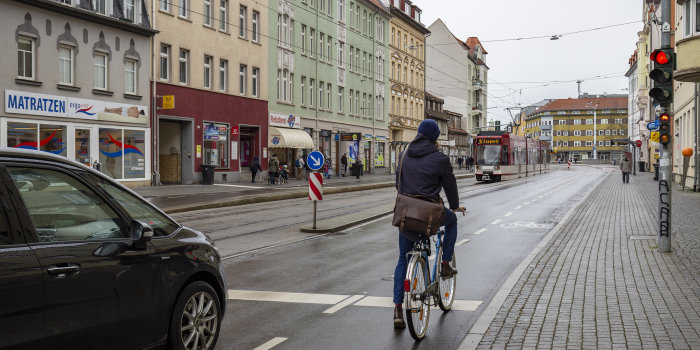 Eine Straße, auf der ein Fahrradfahrer, ein Auto, eine Straßenbahn und ein Fußgänger zu sehen sind. 