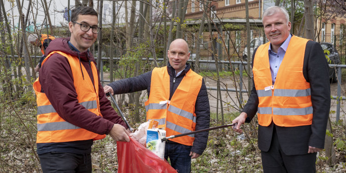 Drei Männer mit orangefarbenen Westen sammeln Müll ein.