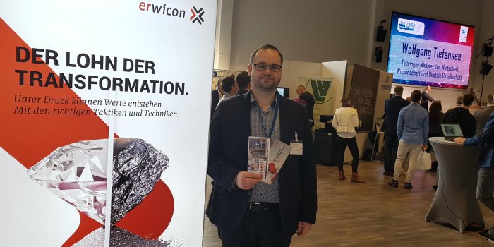 Foto: IT-Leistungsschau 03/2019 © Stadtverwaltung Erfurt Ein Mann steht zwischen einem Werbebanner vom Wirtschaftskongress erwicon und einer Bühne mit Redner und Leinwand.