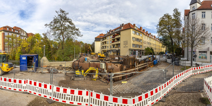 Ein Bauzaun, dahinter ein großes Rohr und Baufahrzeuge