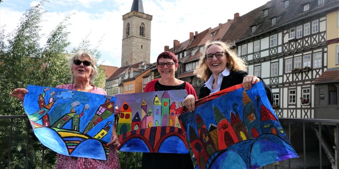 Drei Frauen stehen vor historischen Brückenhäusern und halten Zeichnungen in den Händen.