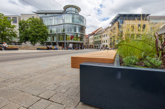mit Bepflanzungen kombinierte Bänke stehen auf breiten Gehwegen entlang einer Straße in der Innenstadt