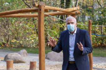 ein Mann mit Mundschutz gestikuliert mit den Händen, im Hintergrund ein Holzgerüst
