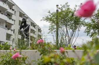 im Vordergrund Blumen, dahinter eine Kunstskulptur mit einem Mehrfamilienhaus im Hintergrund