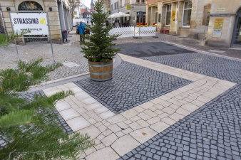 Eine Platz mit Pflastersteinen und eingearbeitet ein Grundriss einer Kirche, in der Mitte ein Tannenbaum im Topf