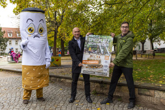 Zwei Männer halten ein Plakat. Neben ihnen steht eine übergrpße Figur in Form einer Zigarette.