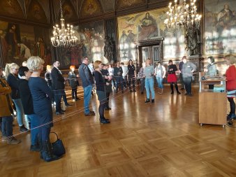 Verschiedenen Menschen stehen in einem Festsaal und wickeln ein Wollknäuel auseinander.