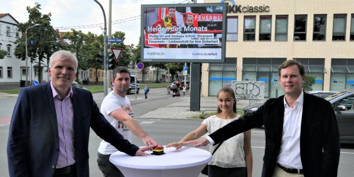 Drei Männer und eine junge Frau stehen um einen Tisch herum und betätigen einen Schaltknopf. 