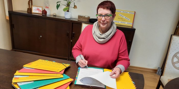 Eine Frau sitzt am Tisch und unterschreibt ein Dokument. Vor ihr liegen viele bunte Mappen.