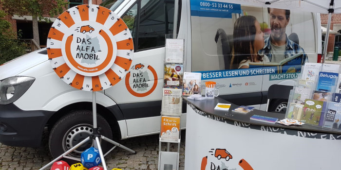 Foto: Alfa-Mobil © Bundesverband Alphabetisierung und Grundbildung e.V. ein Infostand mit einem Transporter im Hintergrund, einem Glücksrad und verschiedenen Infomaterialien