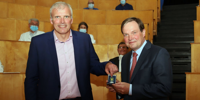 Zwei Männer im Anzug, der Größere übergibt dem Kleineren eine Medaille.