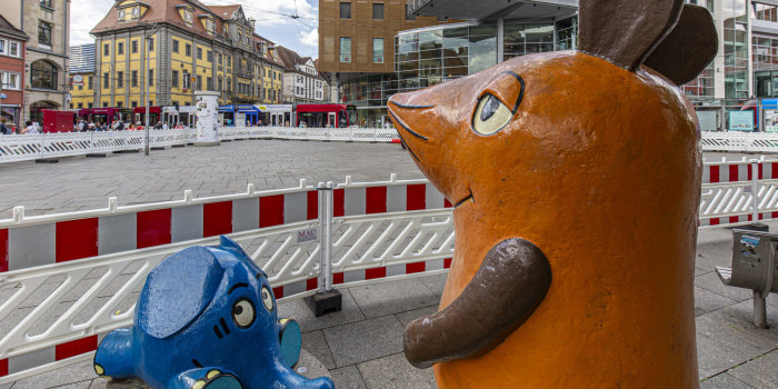 Die Kika-Figuren Maus und Elefant stehen auf einem großen Innenstadtplatz, hinter ihnen sind Bauzäune.