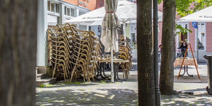 Zwischen einer Reihe junger Bäume in der Stadt stehen übereinander gesteckte Stühle vor einem Restaurant.