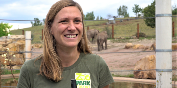 Eine blonde Frau lächelt, im Hintergrund sind Elefanten in einer Anlage zu sehen.