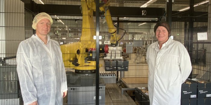 Zwei Männer in Hygienekitteln stehen vor einem eingezäunten Roboterarm in einer Produktionshalle. 