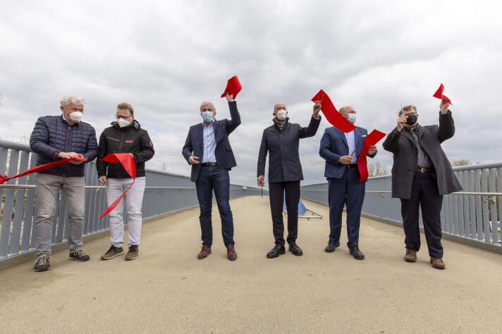 sechs Männer halten auf einer Brücke nach dem Durchschneiden eines roten Bandes die Einzelteile in ihren Händen