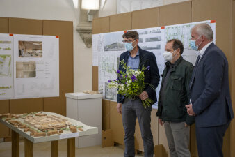 drei Männer stehen vor einem architektonischen Holzmodell, einer hält einen Blumenstrauß in der Hand