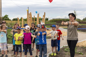 mehrere Kindergartenkinder und eine Frau auf einem Spielplatz, sie halten die Daumen in die Luft