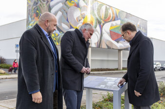 drei Männer stehen an einer Informationstafel, im Hintergrund ein großes Wandmosaik