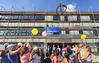 im Vordergrund stehen mehrere Männer vor einem eingerüsteten Rohbau, im Vordergrund lassen Kinder Ballons steigen
