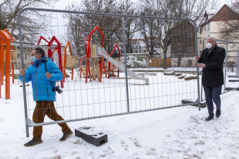 zwei Männer tragen einen Bauzaun, im Hintergrund ein Spielplatz