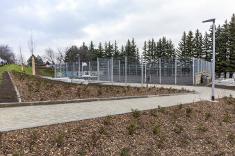 eine Grünfläche, Pflanzflächen im Vordergrund, ein eingezäunter Bolzplatz im Hintergrund