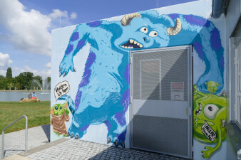 eine mit Graffiti mit Monster-Motiv gestaltete Fassade, im Hintergrund ein Park mit Teich