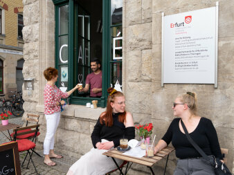 Zwei Frauen sitzen auf Stühlen vor einem Café, ein Mann reicht einer Frau einen To-Go-Becher durch ein Fenster.