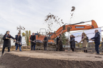 Auf der Baustelle – vor einem orangenen Bagger – versammeln sich die beteiligten Akteure zum Spatenstich