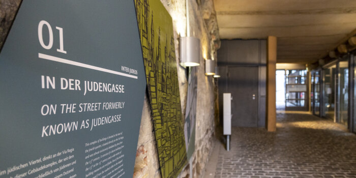 Blick in eine überdachte ganz, an der Mauer links hängt eine Informationstafel mit dem Titel "In der Judengasse"