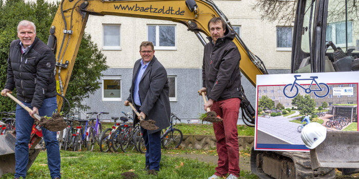 Drei Männer stehen neben einem Bagger. Sie haben jeder einen Spaten in der Hand.