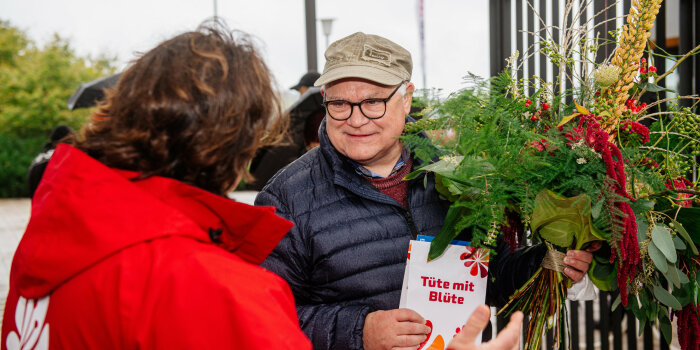 ein Mann mit einem Blumenstrauß und einer Präsenttüte in der Hand spricht mit einer Frau, die ihm gegenübersteht