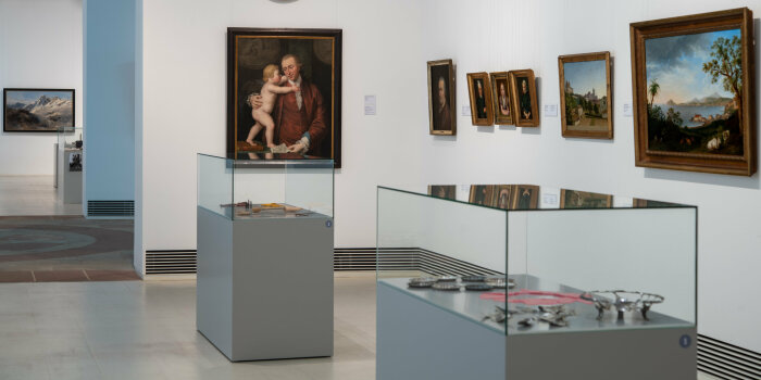 ein Ausstellungraum in einem Museum mit verschiedenen Gemälden an der Wand und im Raum stehenden Vitrinen