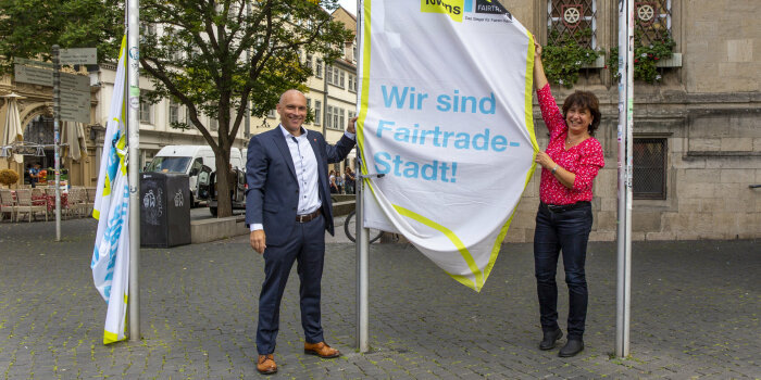ein Mann und eine Frau hissen eine Flagge, auf der steht: "Wir sind Fairtrade-Stadt!"