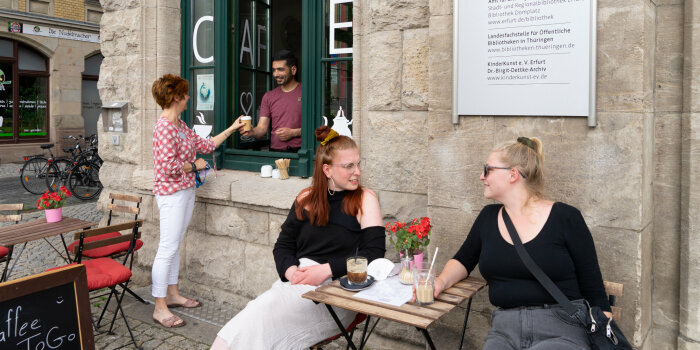 zwei Frauen sitzen auf Stühlen vor einem Café, ein Mann reicht einer Frau einen To-Go-Becher durch ein Fenster