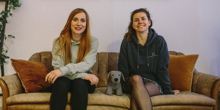 Zwei Frauen sitzen auf einem Sofa. Zwischen ihnen steht ein Kuscheltierhund.