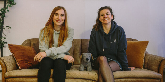 Zwei Frauen sitzen auf einem Sofa. Zwischen ihnen steht ein Stoffhund.