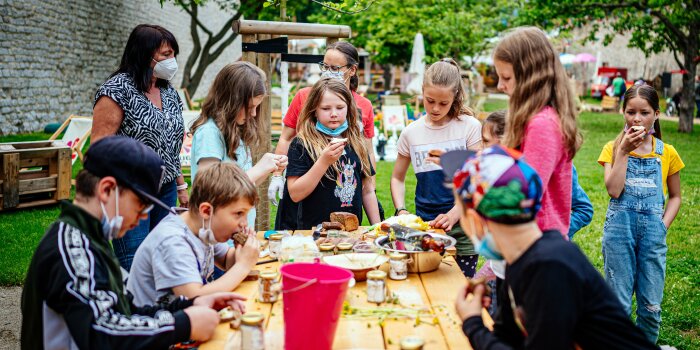 mehrere Kinder stehen im Grünen um einen Tisch herum und essen belegte Brote