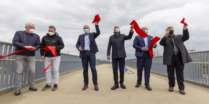sechs Männer halten auf einer Brücke nach dem Durchschneiden eines roten Bandes die Einzelteile in ihren Händen