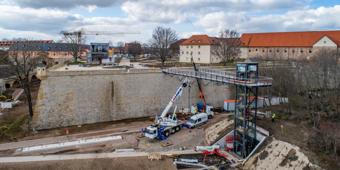 Foto: Die Brücke vom Fahrstuhl zum Plateau © Stadtverwaltung Erfurt Eine Baustelle mit einem Fahrstuhlturm, der über eine Brücke mit einer Festungsmauer verbunden wird.