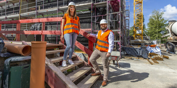 eine Frau und ein Mann mit Warnweste und Helm stehen auf einer Holztreppe auf einer Baustelle
