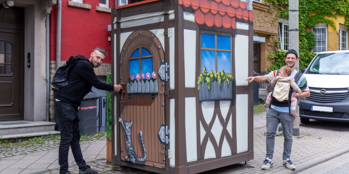 zwei Männer, einer von ihnen trägt ein Baby, an einem mit Graffiti gestalteten Häuschen