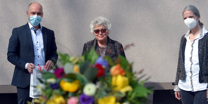 Ein Mann und zwei Frauen stehen nebeneinander, vor ihnen in der Mitte ist ein Blumenstrauß zu sehen. 