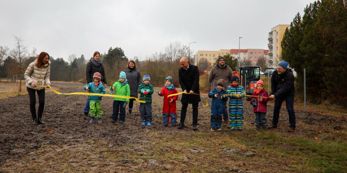 Auf einer Wiese stehen mehrere Erwachsene und Kinder und schneiden ein gelbes Band durch.