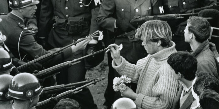 historische Aufnahme eines Mannes, der Blumen in die Läufe von Gewehren steckt, die von Soldaten gehalten werden
