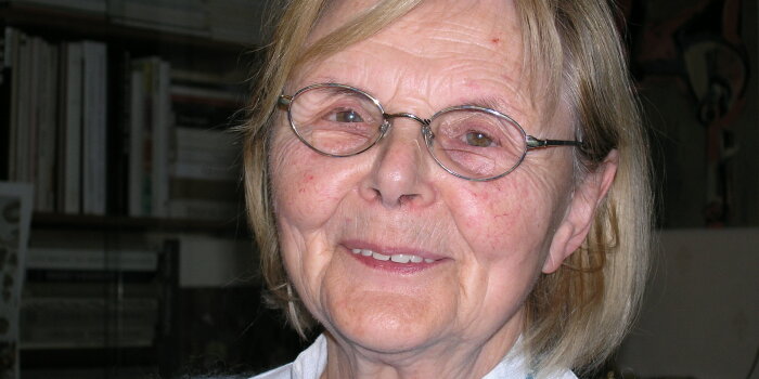 Eine ältere, lächelnde Dame mit blonden Haaren, Brille und blauem Pullover.